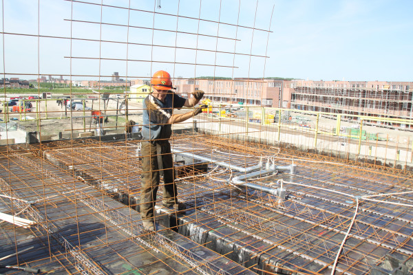 Een bouwvakker met een oranje helm werkt op een bouwplaats aan een betonnen vloer, waarbij hij een metalen rasterconstructie installeert voor gewapend beton.